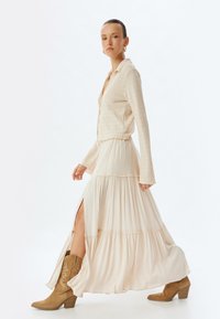 Cardigan texturé beige clair associé à une jupe maxi crème à volants, fluide, avec une fente latérale, complétée par des bottes de cowboy couleur tan.