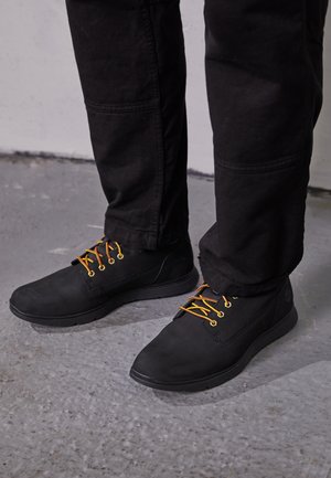 Osoba ubrana w czarne spodnie i czarne casualowe buty z pomarańczowymi sznurówkami stoi na szarej betonowej podłodze.