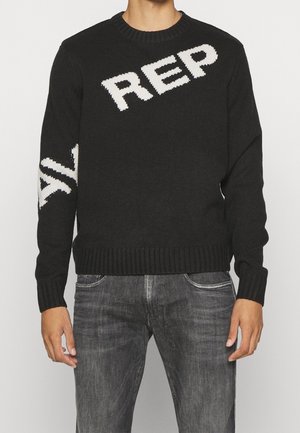 Homme portant un pull en maille noir avec le texte blanc "REP" sur la poitrine et un texte partiellement visible sur la manche gauche, assorti à un jean noir délavé.