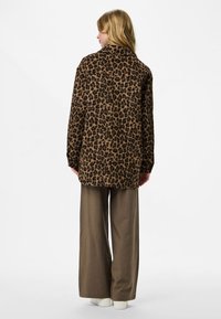 Chaqueta de estampado de leopardo en tela suave, de mangas largas y con un corte relajado, combinada con pantalones marrones de pierna ancha. Zapatos blancos visibles.