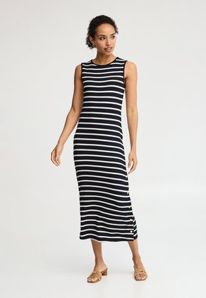 Femme debout portant une robe midi sans manches rayée bleu marine et blanche avec des boutons sur le côté et des sandales à talons couleur beige.