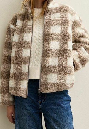 Osoba ubrana w beżową i białą kratowaną kurtkę sherpa na kremowym swetrze w warkocze i niebieskich dżinsach na jednolitym tle.