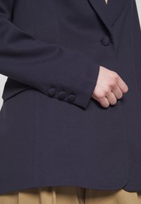 Gros plan sur la main d'une personne ajustant le poignet d'un blazer bleu marine foncé avec des boutons recouverts de tissu, portant un pantalon beige.