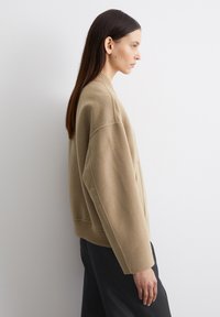 Beige, cropped sweater med en tekstureret finish, ribbet kant og løstsiddende ærmer. Båret over sorte bukser, vist fra en sidevinkel.