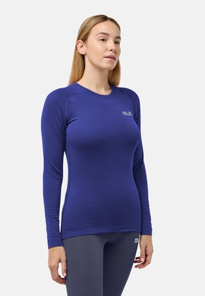 Frau mit glattem blonden Haar, die ein eng anliegendes blaues Langarmshirt und dunkle Leggings trägt, steht vor einem einfarbigen Hintergrund.