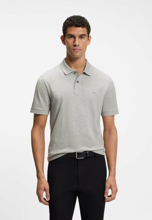 Młody mężczyzna w jasnoszarej koszulce polo z krótkim rękawem wpuszczonej w czarne spodnie z czarnym paskiem, stojący na jednolitym białym tle.