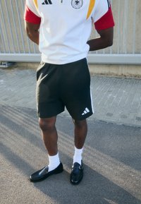 T-shirt branca com fecho éclair e destaques em vermelho e amarelo, calções pretos com riscas brancas na lateral, sapatos pretos e meias brancas. Estilo desportivo.