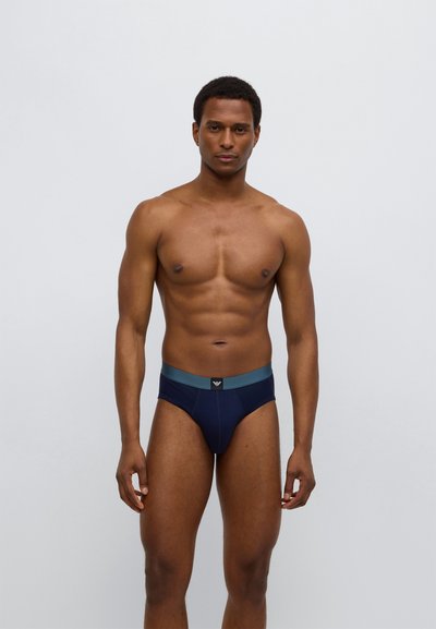 Vyriškos tamsiai mėlynos spalvos trumpikės su švelniu elastiniu liemeniu tamsiai žalsvos spalvos, centre - logotipo ženkliukas. Lygus audinys, aptempantis dizainas.