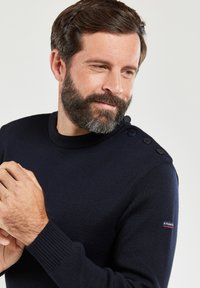 Marineblauer Strickpullover mit einem hohen Rundhalsausschnitt, der über Knopfakzente an der Schulter verfügt. Das Material wirkt weich und strukturiert.