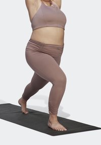 Bruine sportbh en gerimpelde legging van een zachte stof, gedragen door een persoon in een uitvalspose op een donkere yogamat.
