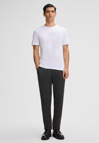 Witte t-shirt met ronde hals, gecombineerd met gestructureerde zwarte broek. Model staat met handen in de zakken en draagt zwarte schoenen. Eenvoudig en aansluitend ontwerp.