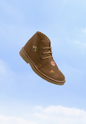 Bottine en daim marron avec broderie florale, bords festonnés et lacets beiges flottant devant un ciel bleu clair.
