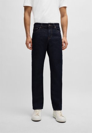 Mężczyzna ubrany w ciemnoniebieskie jeansy o prostych nogawkach, białą koszulkę i białe trampki stoi na jednolitym tle.