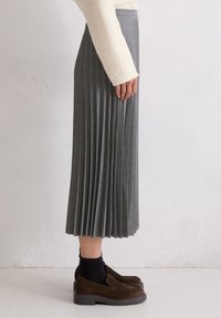 Grijze geplooide rok van stof, met verticale plooien, tot halverwege de kuit en een gladde tailleband. G gedragen met een paar bruine schoenen en zwarte sokken.