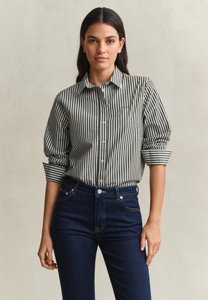 Femme aux cheveux raides et foncés portant une chemise boutonnée à rayures verticales avec manches retroussées et un jean bleu foncé ajusté, debout devant un fond neutre.