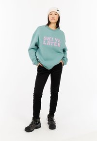 Žena v bielom čiapke, tyrkysovom mikinovom s nápisom "SKI YA LATER", čiernych nohaviciach a turistických topánkach, stojaca so zapnutými rukami v vreckách na bielom pozadí.