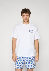 Hvid bomulds t-shirt med rund halsudskæring og korte ærmer, prydet med et blåt og guld logo patch. Sammen med blå mønstrede shorts.
