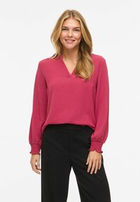 Femme souriante aux longs cheveux blonds portant un chemisier texturé rose profond à col en V avec des poignets smockés et un pantalon noir ample, sur fond blanc.