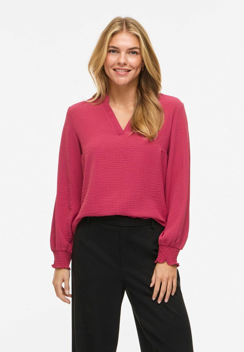 Femme souriante aux longs cheveux blonds portant un chemisier texturé rose profond à col en V avec des poignets smockés et un pantalon noir ample, sur fond blanc.
