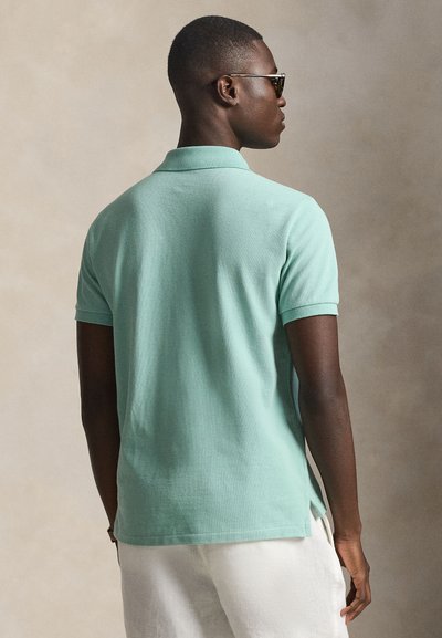 Άνδρας που φοράει ανοιχτοπράσινο κοντομάνικο πόλο και άσπρο παντελόνι, κοιτάζει πλάγια με γυαλιά ηλίου μπροστά σε ουδέτερο φόντο.