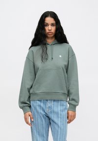 Groene hoodie met een voorzak en geribde manchetten. Heeft subtiele branding op de borst. Gepaard met lichtblauwe gestreepte jeans.