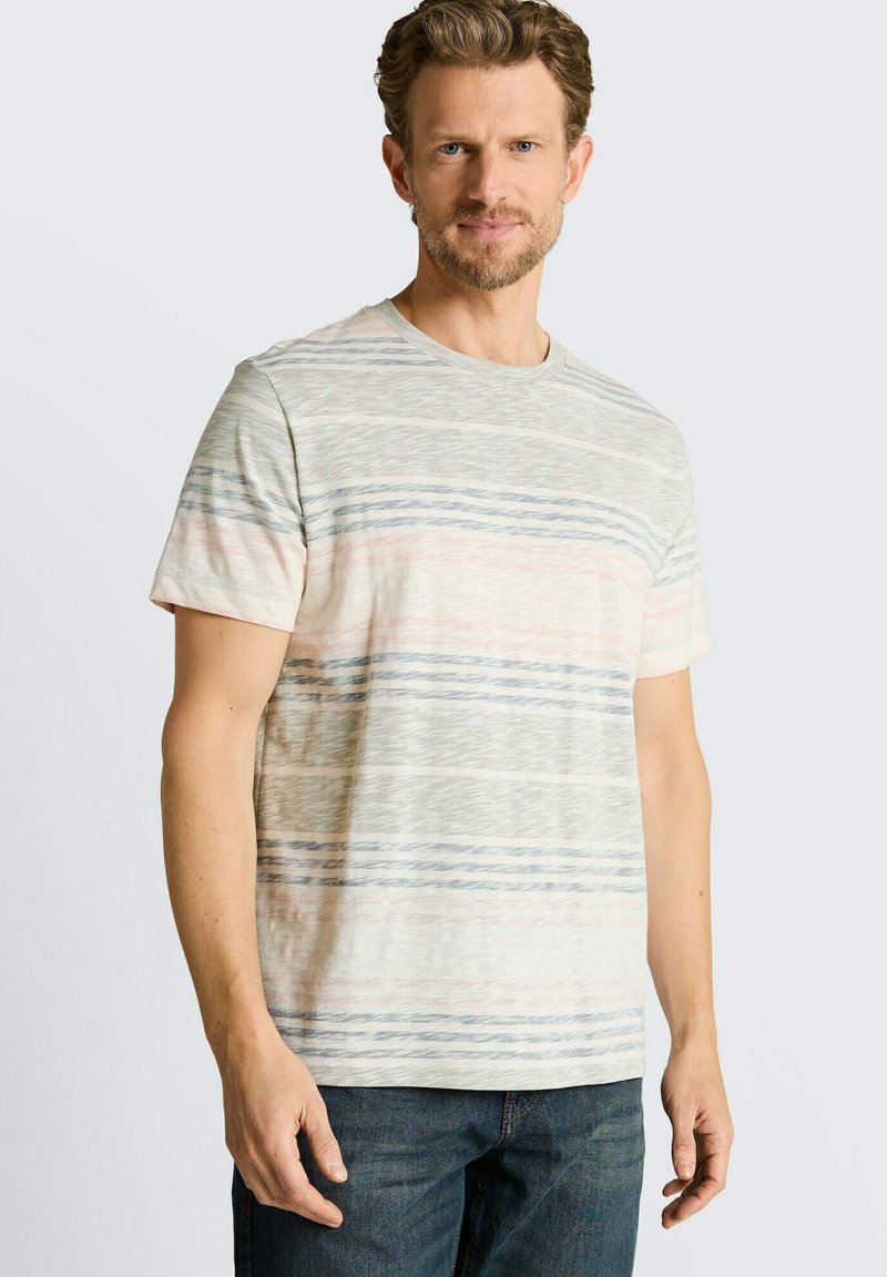 Mann mit kurzen lockigen Haaren und Bart, der ein beige kurzärmeliges Hemd mit horizontalen grauen und pinken Streifen und blaue Jeans trägt.
