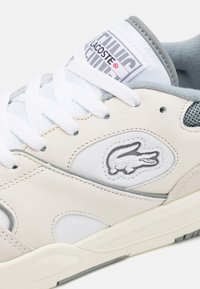 Baskets blanches et grises avec une tige en cuir et textile, un bout rond, des lacets blancs et un logo crocodile embossé bien visible.