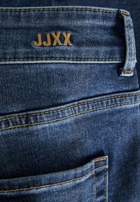 Mėlynos džinsinės kelnės su matoma užpakaline kišene, kontrastinga geltona siūle ir siuvinėtu ženkliuku "JJXX" ant juosmens.