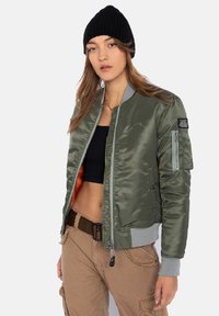 Olivgrüne Bomberjacke mit satiniertem Finish, grauen Rippbündchen und einer Reißverschlusstasche am linken Ärmel. Das Innere ist mit orangefarbenem Futter ausgestattet.