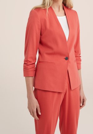 Kobieta w koralowej, dopasowanej marynarce z jednym czarnym guzikiem oraz dopasowanych spodniach z szerokimi nogawkami, biała bluzka pod spodem, stoi na jednolitym tle.