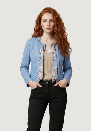 Giovane donna con lunghi capelli ricci rossi che indossa una giacca blu sopra una maglia beige e pantaloni neri, in piedi con le mani nelle tasche.