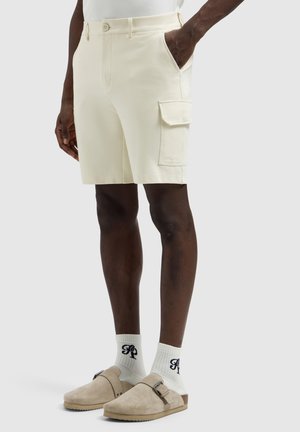 Man draagt crème-kleurige cargoshorts met zijzakken, witte sokken met zwarte letters en beige instapschoenen met gespen.