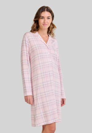 Mujer con una camisa de noche de cuadros rosa hasta la rodilla, con mangas largas y botones, de pie frente a un fondo gris claro liso.