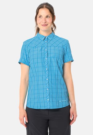 Frau, die ein kurzärmliges blau kariertes Hemd mit Knöpfen und schwarze Hosen trägt, steht vor einem einfarbigen hellen Hintergrund.