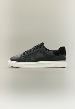Baskets basses en cuir noir avec lacets noirs et semelle blanche, montrées de profil sur un fond gris neutre.