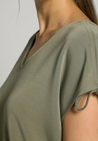 Top de manga corta en verde oliva, fabricado en un tejido suave, con escote en V y detalle fruncido en las aberturas de las mangas. Textura lisa, corte relajado.