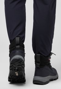 Scarpe da trekking nere con tomaia testurizzata, suole grigie e etichetta da tirare. Indossate con pantaloni scuri e aderenti. Inquadratura dal basso che mette a fuoco la calzatura.