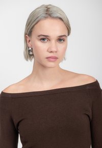 Brauner, schulterfreier Strickpullover mit weicher Textur. Das Model trägt silberne, runde Ohrringe und hat hellblonde Haare. Neutraler Hintergrund.