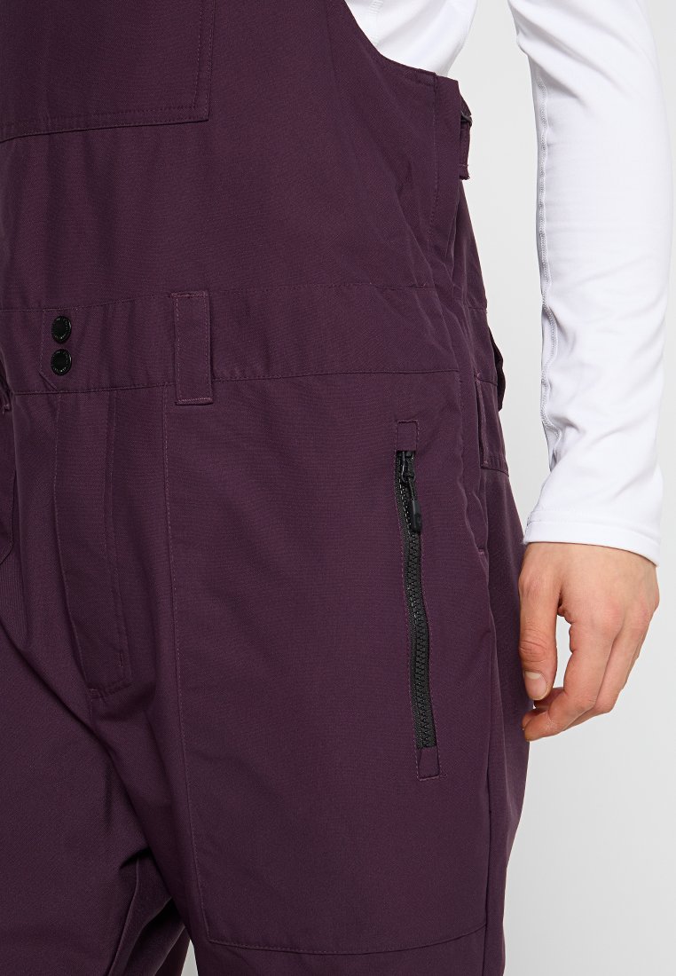 Fioletowe ogrodniczki z zamkiem błyskawicznym na boku, z czarnymi guzikami i teksturowanym materiałem, część warstwowej stylizacji z białą koszulą z długim rękawem.