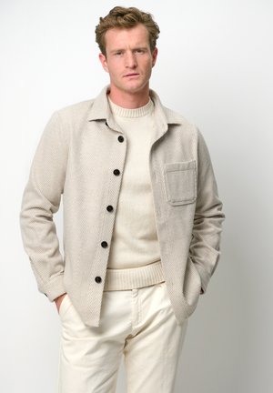 Beige jasje met een visgraatpatroon, met een kraag, knoopsluiting en een borstzak, gedragen over een crèmekleurige trui en lichte broek.