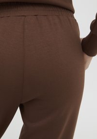 Bruna stretchbyxor med elastisk midja, med en slät yta och figursydd design, visade från baksidan.