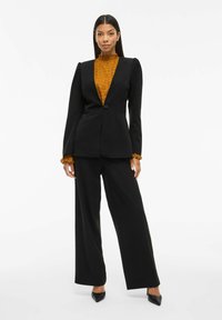 Černý blazer s jedním knoflíkem, spárovaný s volnými černými kalhotami. Pod ním je strukturovaná hořčicová blůza s volánkovými manžetami.