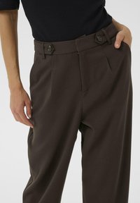 Pantaloni eleganți maro cu buzunare laterale, față plisată și un tiv lat cu două accente de buton. Textură de țesătură netedă.