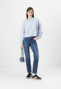 Lys blå kortermet genser, ribbet kant, kombinert med blå jeans. Modellen har på seg svarte sneakers med hvite striper og holder en liten blå veske.