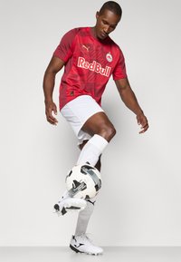 Maillot de football rouge avec motif graphique et logo "Red Bull", associé à un short blanc et un ballon de soccer noir et blanc. Joueur équilibrant le ballon sur son pied.