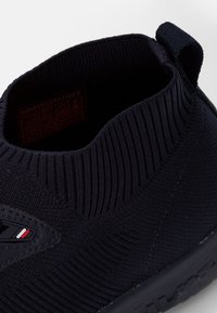 Gros plan sur une basket à enfiler noire en tricot avec col côtelé, languette de tirage et petit logo Tommy Hilfiger sur le côté.