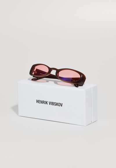 Ochelari de soare rectangulari, maro, cu lentile roz, așezați pe o cutie albă etichetată "HENRIK VIBSKOV." Rama are un finisaj elegant și neted.