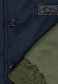 Marineblå jakke i stof med en grøn ærme. Har runde sorte knapper og en rektangulær label med bjerggrafik. Ribbet grøn manchet.