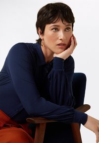 Marinblå långärmad skjorta med knapppdetaljer, avslappnad passform, textil med struktur, tillsammans med en rostfärgad nederdel. Modell sitter på en trästol.