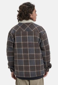 Camisa chaqueta de polar a cuadros marrón y gris con un collar color crema, textura suave y puños ajustados, que presenta una silueta amplia.